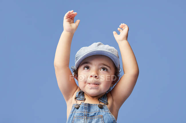 Charming barefoot child in denim dress and dress with curly hair looking up with arms raised while dancing on blue background — Stock Photo