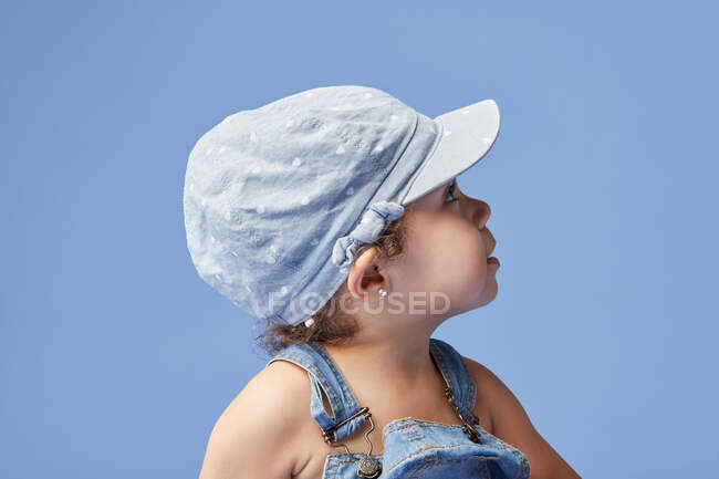 Vue latérale de charmant enfant en robe de denim et avec les cheveux bouclés regardant loin tout en se tenant sur fond bleu — Photo de stock