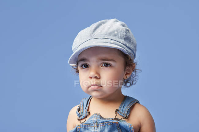 Adorable petit enfant bouleversé aux yeux marron portant des vêtements décontractés et un chapeau sur fond bleu détournant les yeux — Photo de stock
