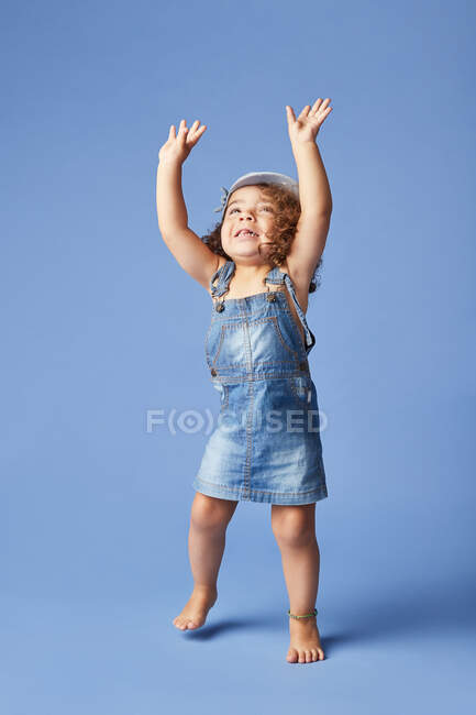 Charming barefoot child in denim dress and hat with curly hair looking up with arms raised while dancing on blue background — Stock Photo