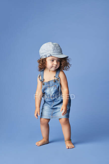Criança descalça encantadora em vestido de ganga e chapéu com cabelo encaracolado olhando para longe enquanto dança no fundo azul — Fotografia de Stock