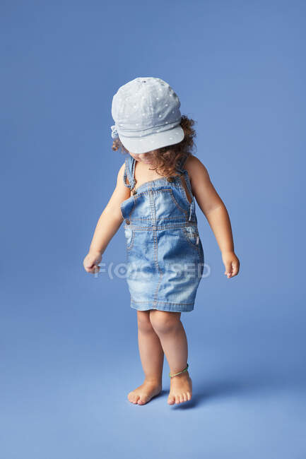 Charmantes barfüßiges Kind in Jeanskleid und Hut mit lockigem Haar, das beim Tanzen auf blauem Hintergrund wegschaut — Stockfoto