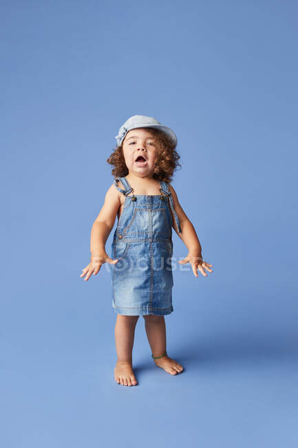 Charmant fröhliches Barfußkind in Jeanskleid und Hut mit lockigem Haar, das in die Kamera schaut, während es auf blauem Hintergrund tanzt — Stockfoto