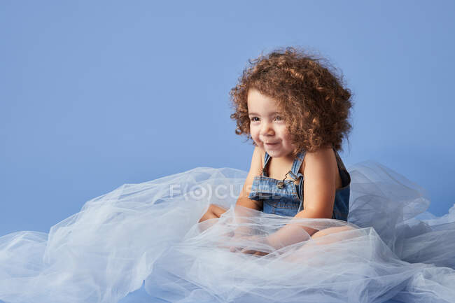 Adorável encaracolado menina sorrindo bonito sentado em tecido fino contra fundo azul e olhando para longe — Fotografia de Stock