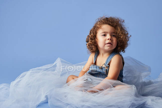 Adorável encaracolado menina sorrindo bonito sentado em tecido fino contra fundo azul e olhando para longe — Fotografia de Stock