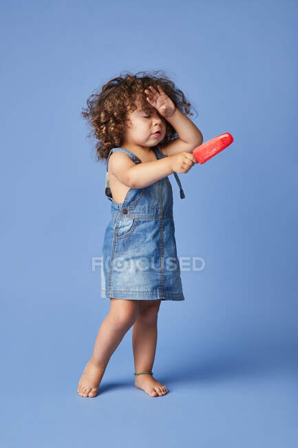 Cuerpo completo de niña molesta con los ojos cerrados manteniendo la mano en la cabeza de pie con helado sobre fondo azul - foto de stock
