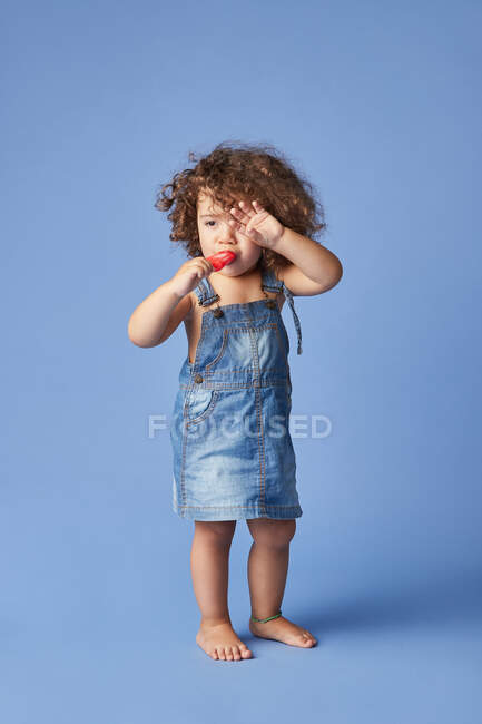 Full body of upset little girl on summer clothes barefooted standing with ice cream against studio blue background — Stock Photo