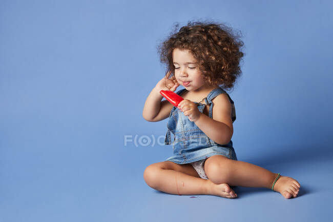 Full body of delighted little girl sitting with melting popsicle against blue background — Stock Photo