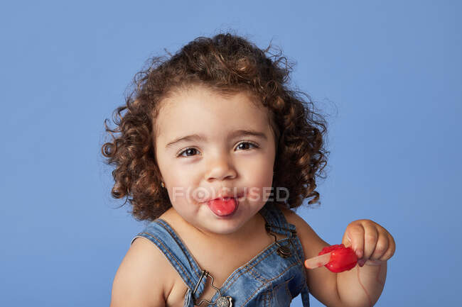 Дивна дівчина в денімському одязі з кучерявим волоссям, що показує язик, споживаючи солодке морозиво на синьому тлі. — стокове фото