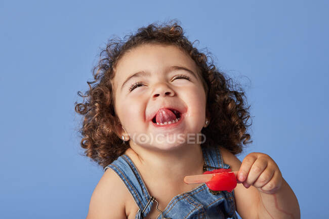 Menina engraçada em roupa de ganga com cabelo encaracolado mostrando língua enquanto come sorvete doce contra fundo azul — Fotografia de Stock