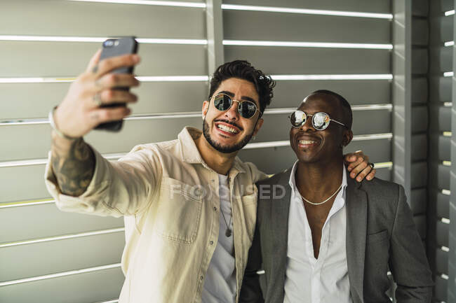 Hombre étnico barbudo feliz con tatuaje abrazando socio negro en traje elegante y gafas de sol mientras toma autorretrato en el teléfono celular - foto de stock