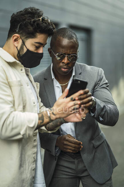 Стиль афроамериканського чоловічого костюма біля етнічного партнера в масці дивитися мобільний телефон на вулицях міста — стокове фото