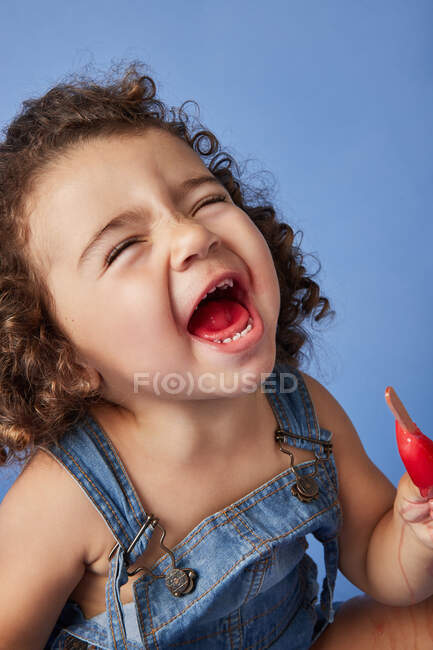 Ragazza divertente in denim vestito con i capelli ricci ridere mentre si mangia gelato dolce sullo sfondo blu — Foto stock