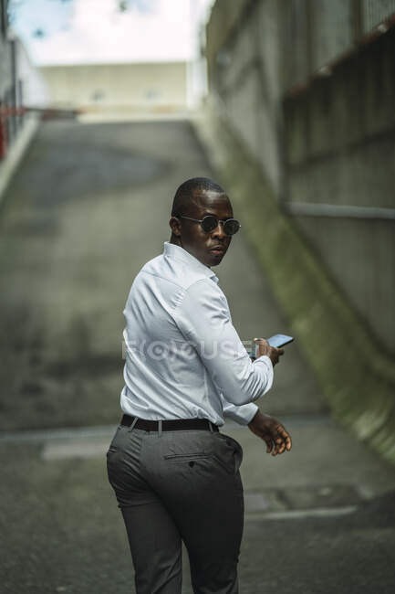 Повернення до афроамериканського чоловіка-підприємця у формальному одязі та сонцезахисних окулярах з мобільним телефоном, що дивиться через плече в місті. — стокове фото