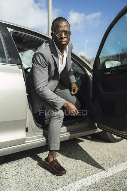Adulto chefe masculino afro-americano em terno elegante e óculos de sol olhando para a câmera do automóvel contemporâneo à luz do sol — Fotografia de Stock