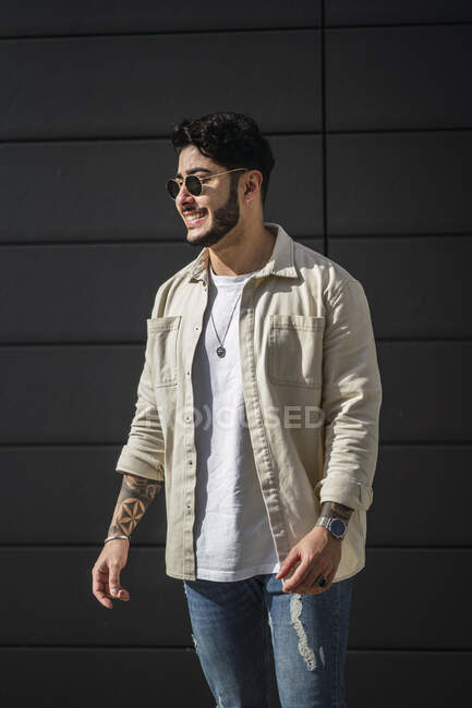 Young content ethnic male with tattoos in ripped jeans and casual shirt looking away on city street — Stock Photo