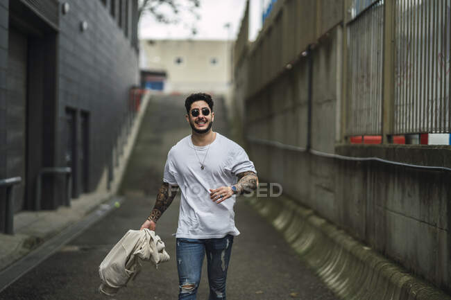 Молодой счастливый татуированный этнический мужчина в солнцезащитных очках и разорванных джинсах, прогуливающийся по дорожке между городскими зданиями — стоковое фото