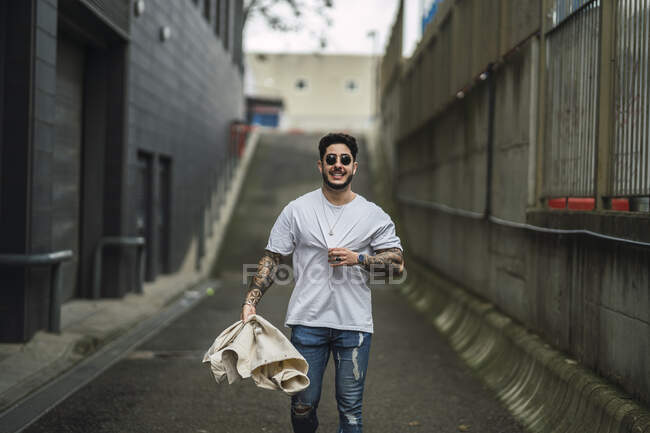 Молодой счастливый татуированный этнический мужчина в солнцезащитных очках и разорванных джинсах, прогуливающийся по дорожке между городскими зданиями — стоковое фото
