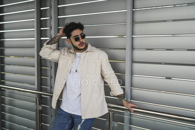 Jovem conteúdo étnico masculino com tatuagens em jeans rasgados e camisa casual olhando para longe na rua da cidade — Fotografia de Stock