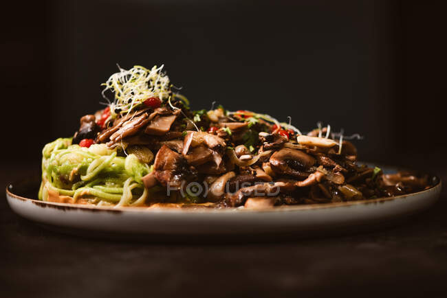 Baixo ângulo de prato vegan gostoso com espaguete de abobrinha e fatias de cogumelos refogados cobertos com bagas vermelhas e brotos de alfafa no fundo escuro — Fotografia de Stock