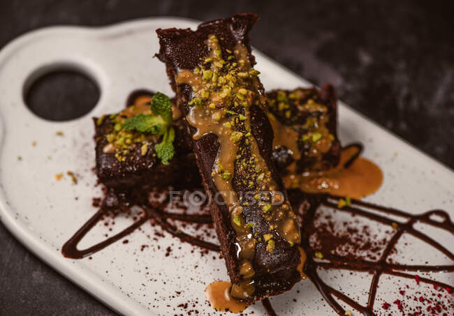 Des brownies savoureux recouverts de beurre d'arachide et de pistaches concassées croquantes près d'une cuillère à glace sur une assiette avec sauce au chocolat — Photo de stock