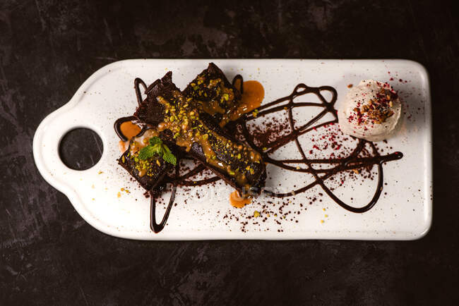 Des brownies savoureux recouverts de beurre d'arachide et de pistaches concassées croquantes près d'une cuillère à glace sur une assiette avec sauce au chocolat — Photo de stock