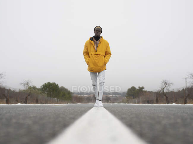 Cuerpo completo a nivel del suelo de hombre afroamericano con las manos en los bolsillos de pie en la carretera de asfalto entre árboles sin hojas y nieve - foto de stock