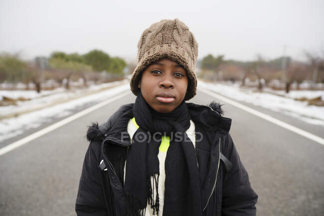 Adolescente niño vistiendo chaqueta caliente bufanda y sombrero de pie en camino de asfalto contra vistosas plantas sin hojas y mirando a la cámara - foto de stock