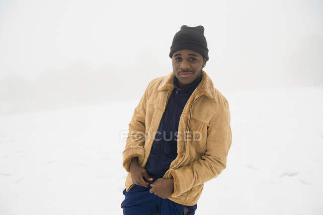 Joven afroamericano usando ropa de invierno de pie en terreno nevado y mirando a la cámara - foto de stock