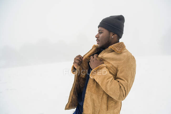 Vista lateral de un joven afroamericano con ropa de invierno de pie en terreno nevado y mirando hacia otro lado - foto de stock