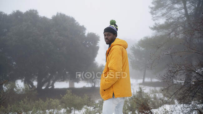 Side view of thoughtful African American man with hands and pockets wearing outerwear standing in snowy park — Stock Photo