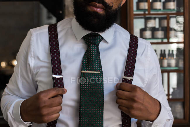Crop unrecognizable ethnic boss wearing elegant white shirt and suspenders putting on tie lip while preparing for work — Foto stock
