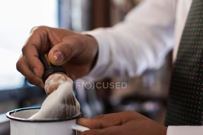 Crop anonymous barber putting brush for shaving into cup while working in barbershop - foto de stock