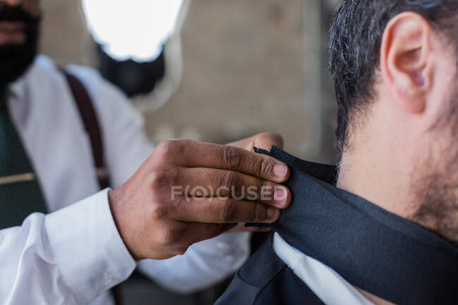 Cropped unrecognizable ethnic male beauty master putting on cape protection on client in hairdressing salon — Stock Photo