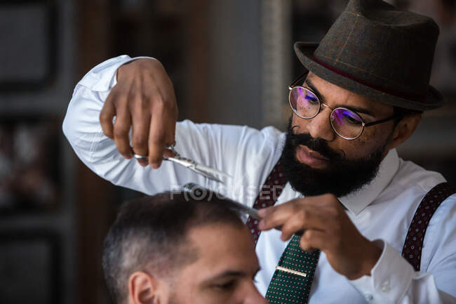 Serious mature bearded ethnic male barber trimming hair of client with scissors during grooming procedure in beauty salon — Foto stock