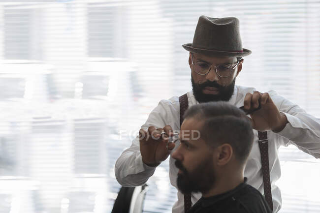 Ernste ältere bärtige männliche ethnische Friseur schneiden Haare der Klientin mit der Schere während der Pflege Verfahren in Schönheitssalon — Stockfoto