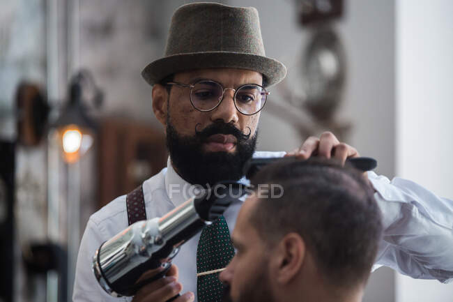 Professional mature ethnic male beauty master drying hair of crop male client using hairdryer in barbershop — Fotografia de Stock