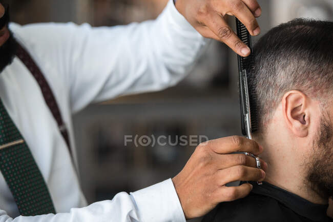 Rogné méconnaissable ethnique homme coiffeur couper les cheveux du client avec des ciseaux pendant la procédure de toilettage dans le salon de beauté — Photo de stock