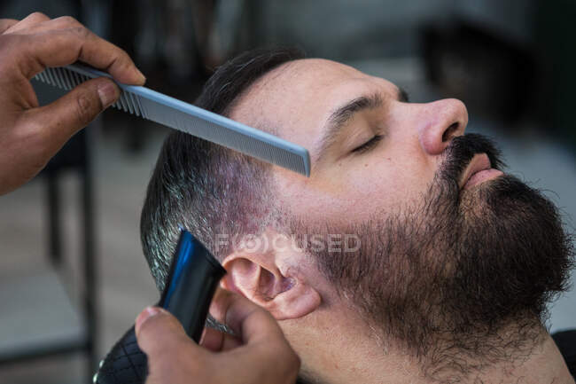 Crop anonymous ethnic male beauty master cutting mustache of bearded client using trimmer and comb in barbershop — Fotografia de Stock