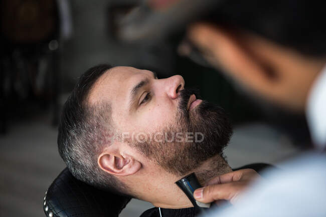 From above crop anonymous ethnic male beauty master cutting mustache of bearded client using trimmer and comb in barbershop — Foto stock