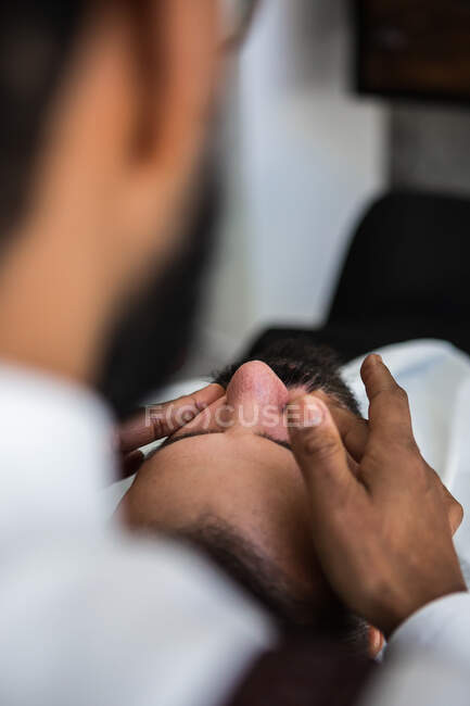 Crop anonymous ethnic male hairdresser applying beauty product on face of man with closed eyes while massaging in barbershop — Stock Photo