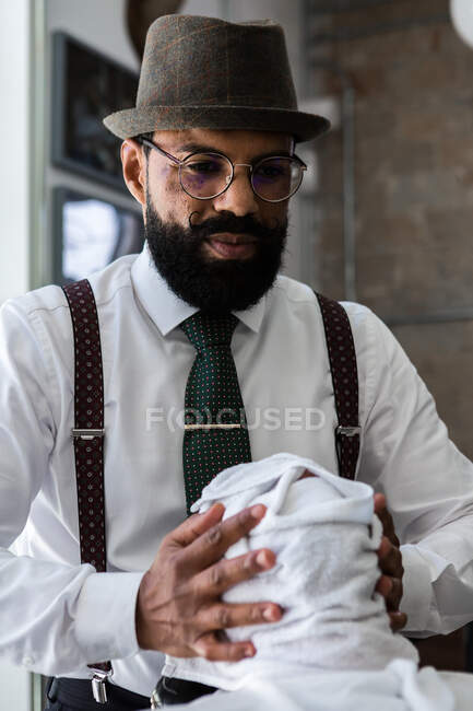 Stylish mature ethnic male stylist in hat and eyewear putting soft wet towel on face of anonymous client in barbershop — Foto stock