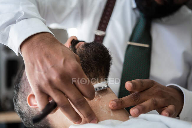 Crop anonymous stylish ethnic male beauty master removing beard of masculine client using straight razor in hairdressing salon — Fotografia de Stock
