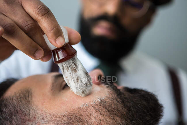 Closeup of crop masculine ethnic beauty master applying smooth shaving soap on face of bearded man using brush in hairdressing salon — Foto stock