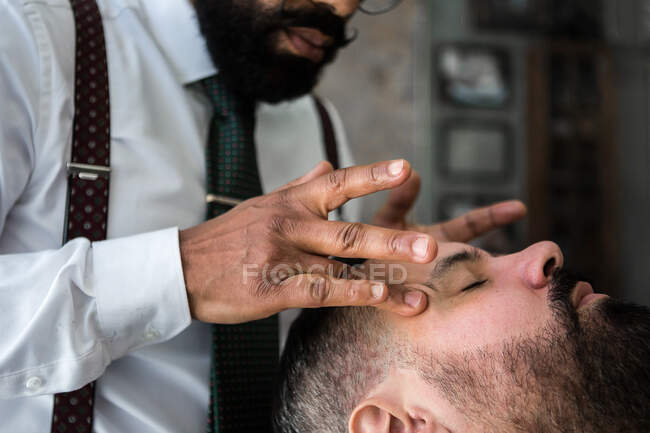 Crop anonymous ethnic male hairdresser applying beauty product on temple of man with closed eyes while massaging face in barbershop — Foto stock