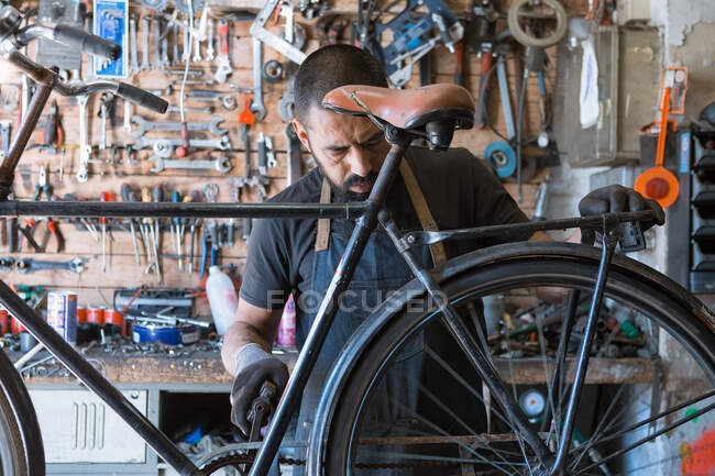 Mecánico masculino concentrado con barba y tatuajes en guantes reparando bicicleta en taller moderno - foto de stock