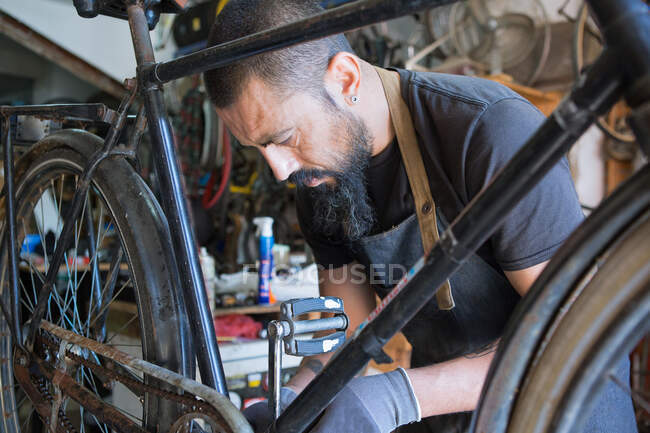 Mecánico masculino concentrado con barba y tatuajes en guantes reparando bicicleta en taller moderno - foto de stock