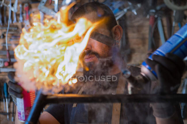 Homem barbudo grave em avental e luvas de proteção usando equipamentos profissionais com fogo para reparação de bicicletas na oficina — Fotografia de Stock