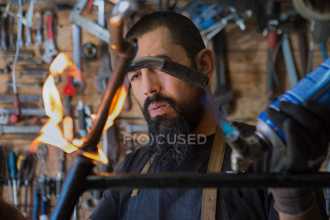 Serious bearded man in apron and protective gloves using professional equipment with fire for repair bicycle in workshop — Stock Photo