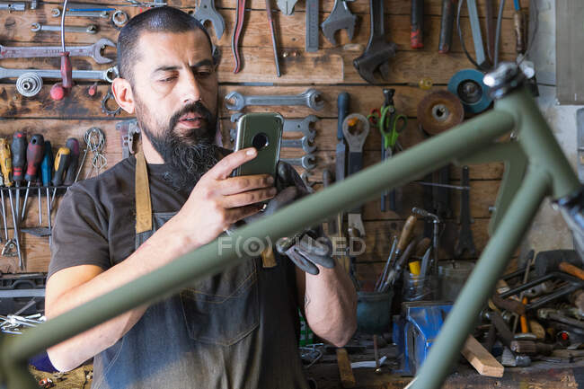 Maestro barbudo concentrado en delantal tomando fotos de bicicleta durante la reparación en garaje - foto de stock